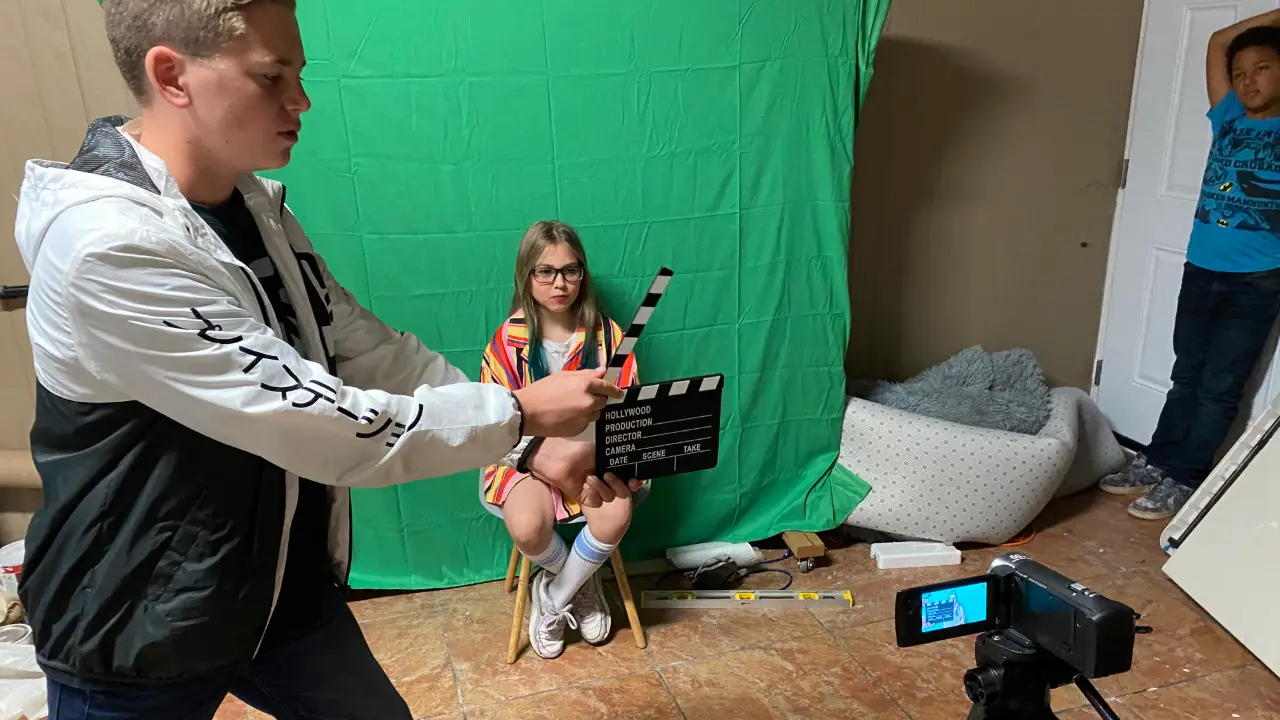 A camper marking a take with a clapperboard while another acts on the green screen at film camp
