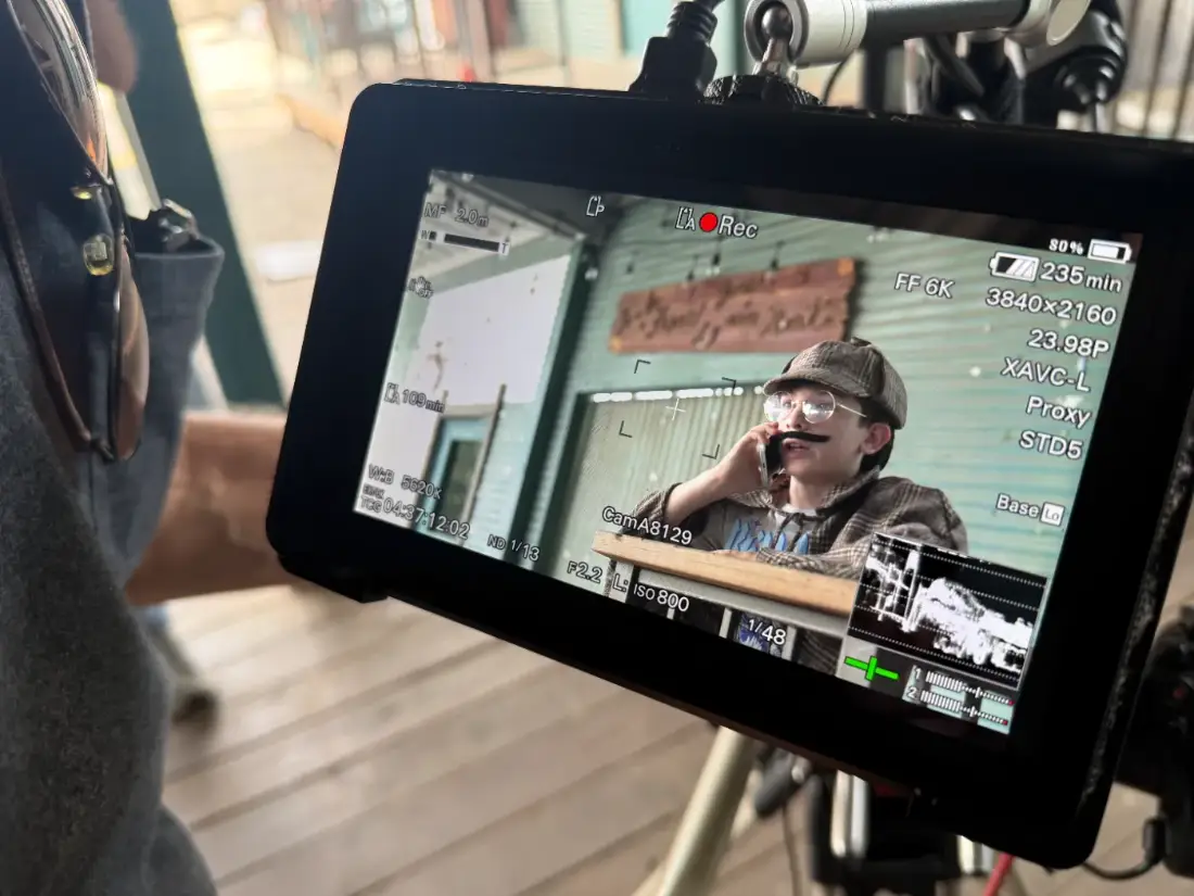 A young actor in a flat cap and moustache on a phone in character, framed up on the camera's monitor mid-take